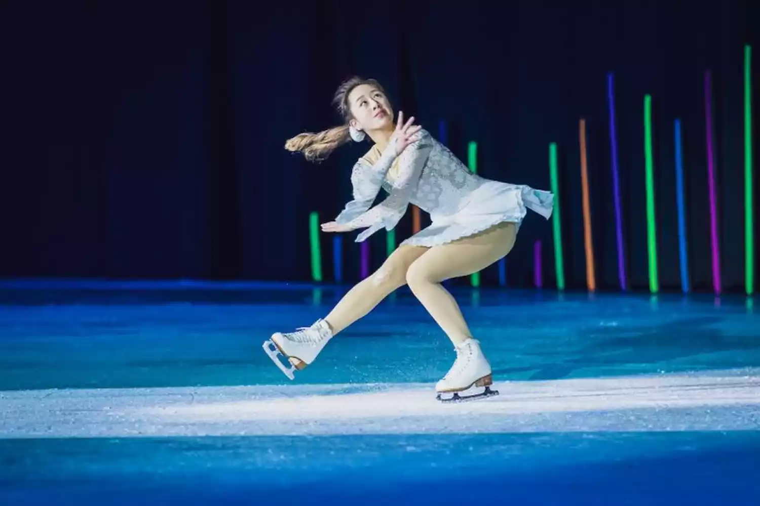 Female figure skater in a white costume performs on ice, with colorful lights in the background.