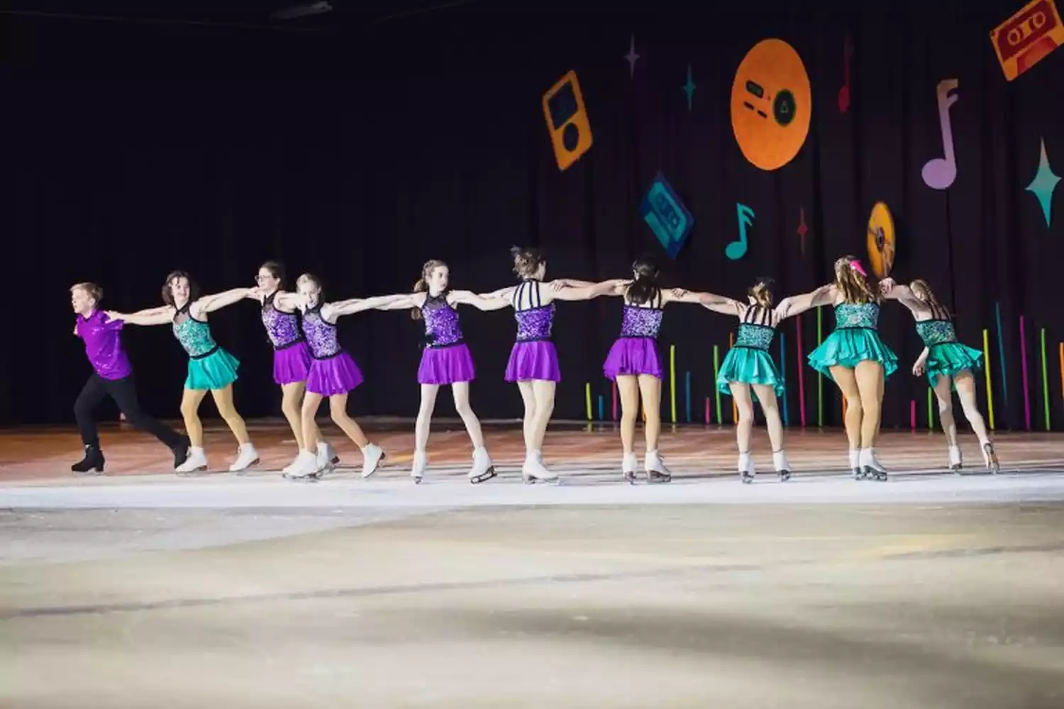A line of young ice skaters in colorful costumes holding hands and skating across an ice rink. The backdrop features retro music-themed decorations.