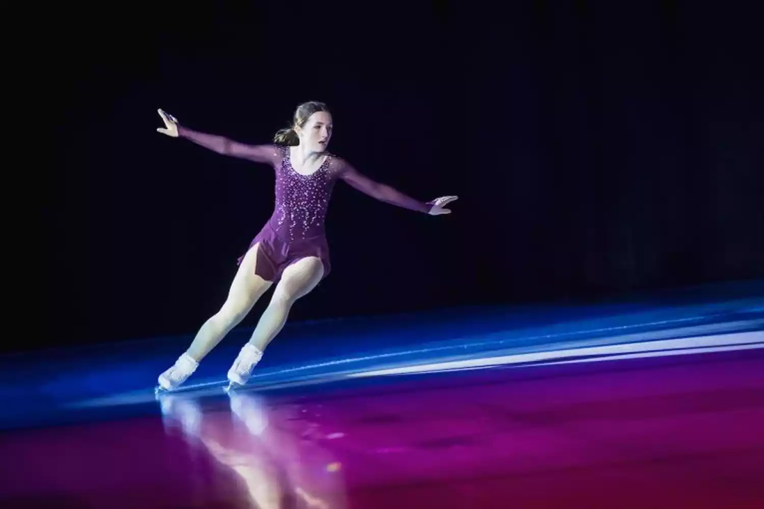Figure skater in a sparkly purple costume performs on an ice rink under dramatic lighting.
