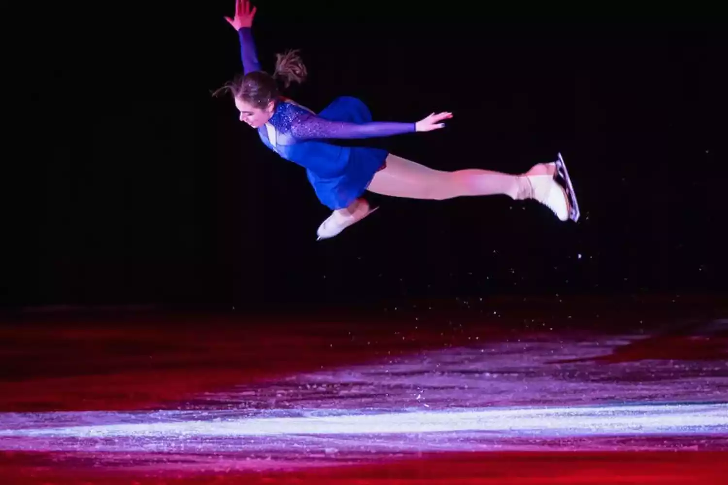 A female figure skater in a blue and purple costume performs a jump on an ice rink with red and purple lighting.