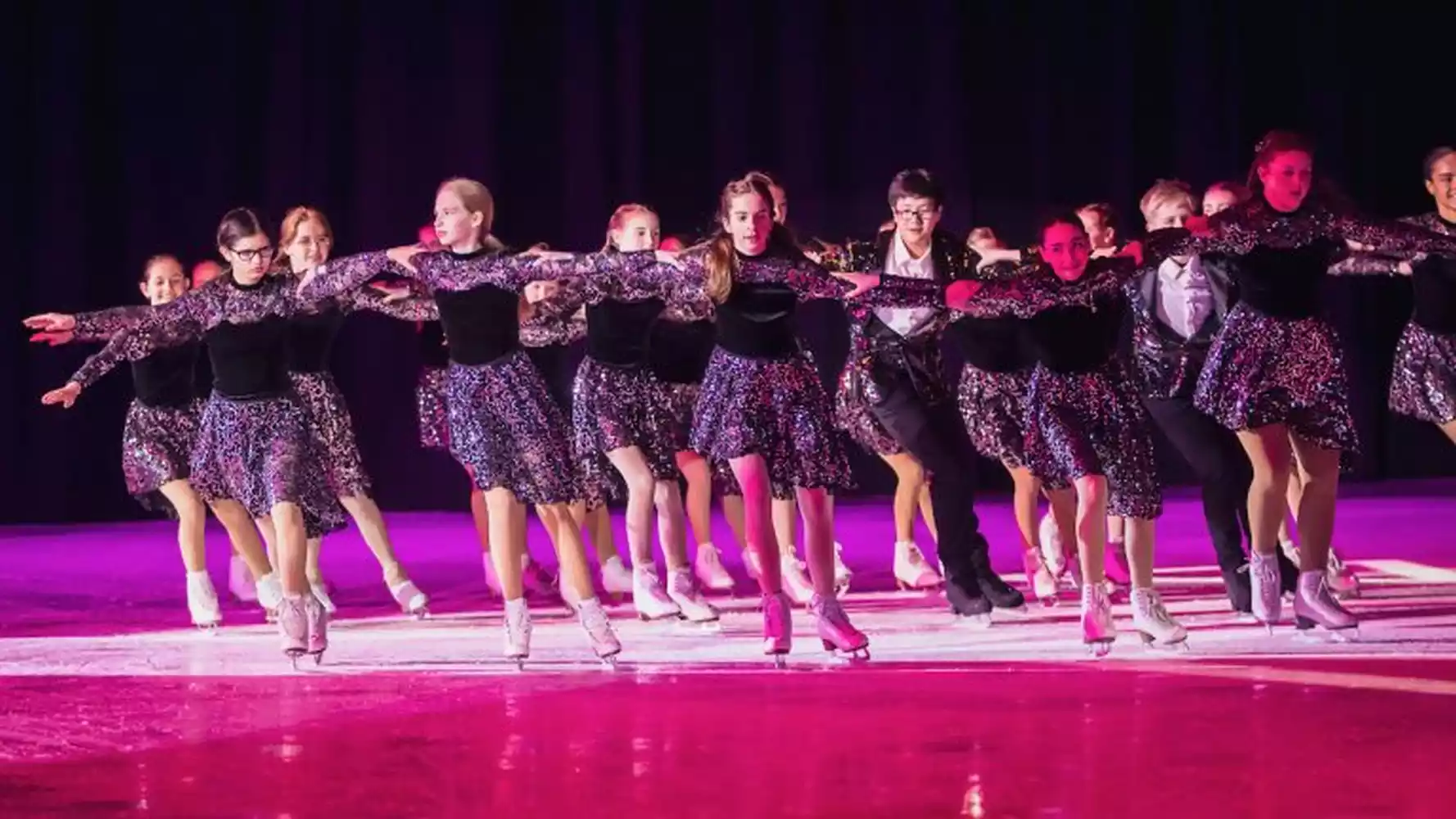 A group of young ice skaters in matching sparkly purple and black costumes perform on an ice rink under purple stage lighting.