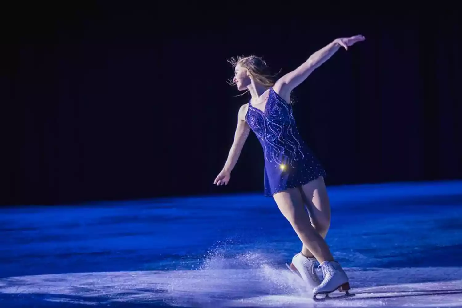 A female figure skater in a sparkling blue dress performs on ice, with arms extended and skates kicking up spray.