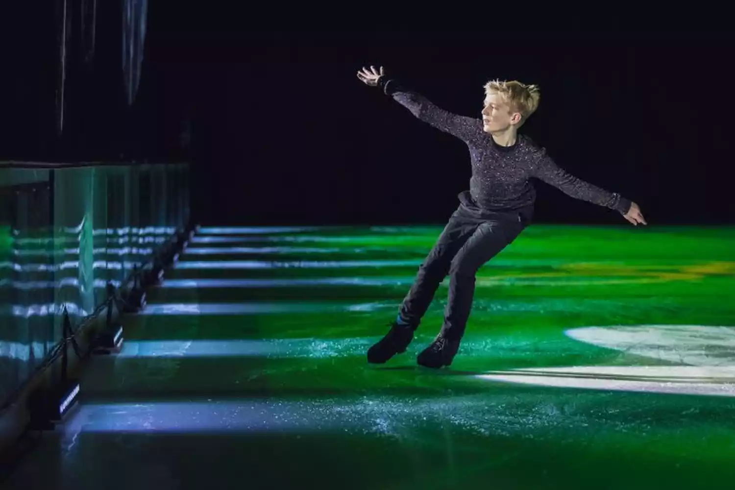 A young male figure skater performs on an ice rink under dramatic green and blue lighting. He is in motion, with one arm extended.