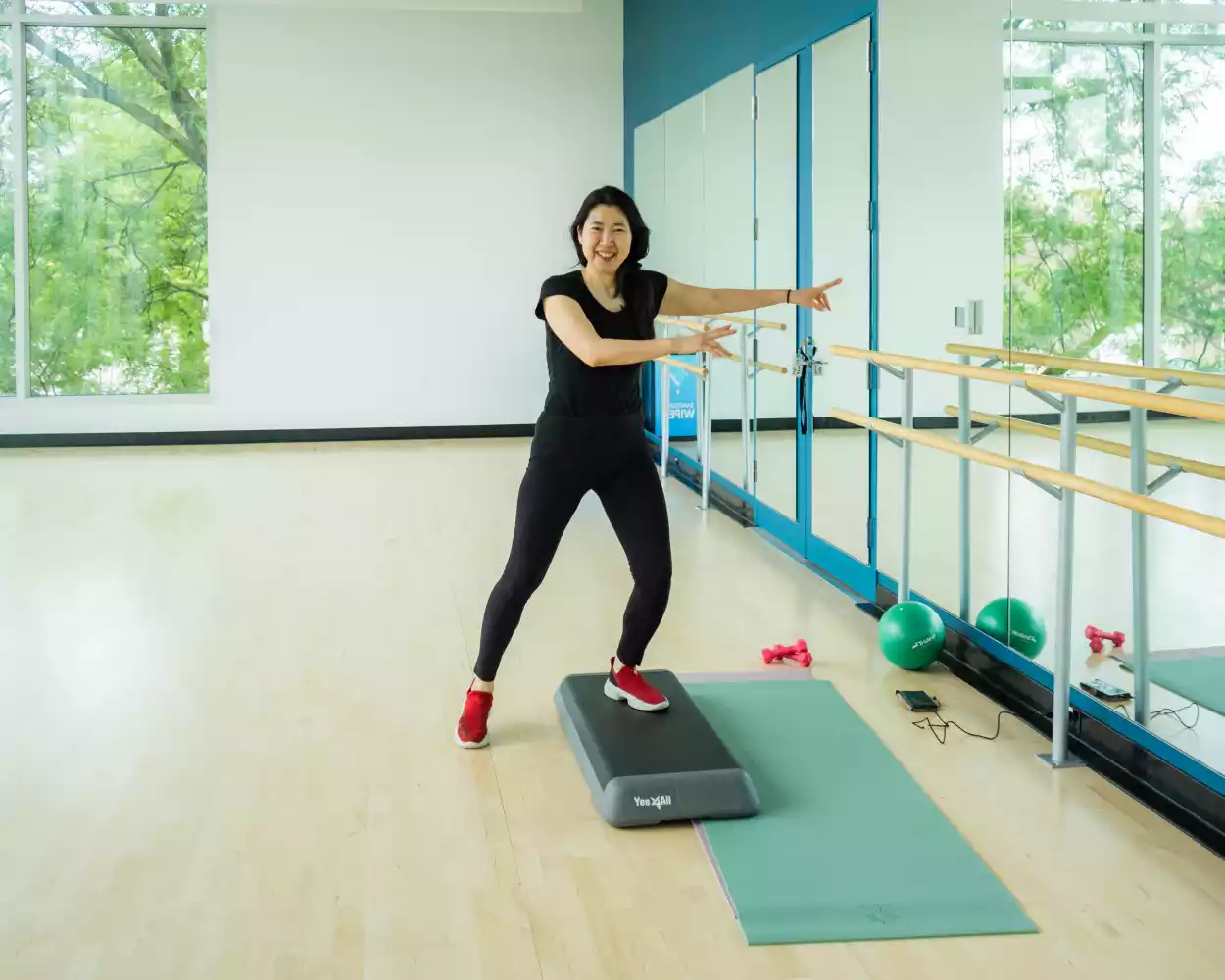 A woman in athletic wear smiles while demonstrating a dance move on a step platform in a fitness studio.