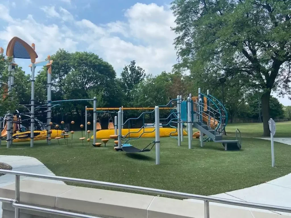 Modern playground equipment with yellow slides, blue accents, and climbing structures on artificial turf, surrounded by trees.