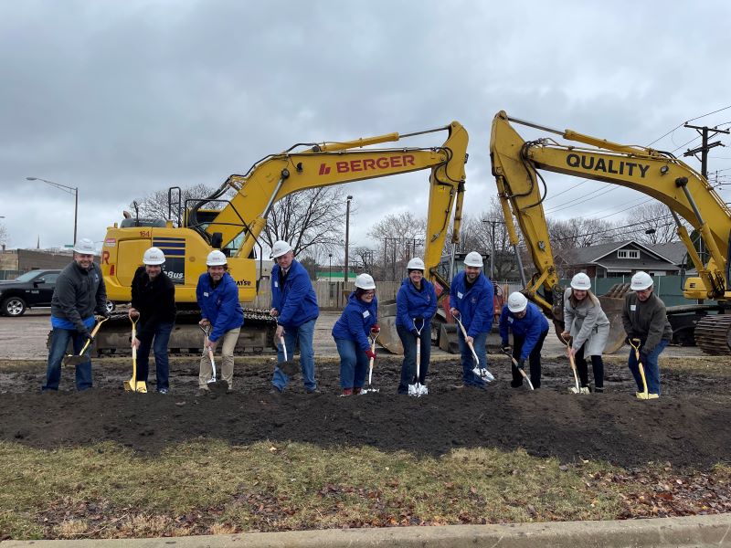A group of nine people in hard hats and blue jackets participate in a groundbreaking ceremony, holding shovels in dirt with two yellow excavators