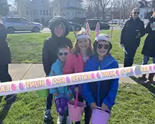 Four children wearing Easter bunny ears and holding baskets stand behind a banner that reads "EASTER EGG HUNT".