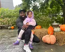 Man in a black jacket and cap holds a young girl in a purple hoodie and leggings. They are posing outdoors with pumpkins and hay bales.