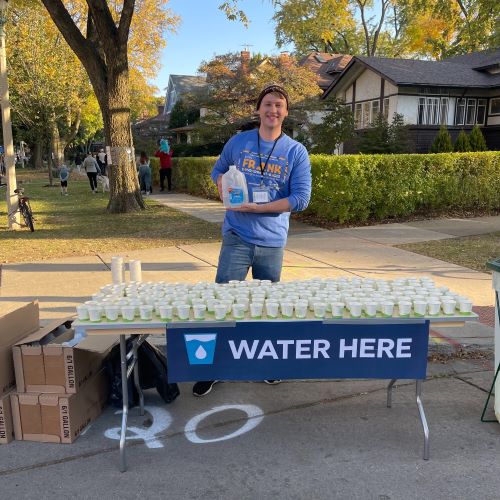 A smiling volunteer stands behind a table laden with small cups of water and a large jug. A banner on the table reads "WATER HERE".