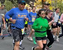 Participants in a race, including a man in a blue shirt with "FRANK LLOYD WRIGHT" and a woman in a green shirt, running together.