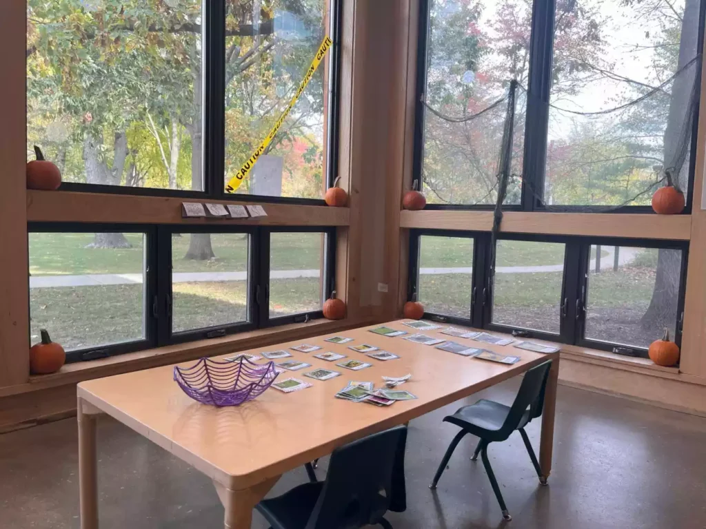 A table with cards laid out, a purple spiderweb basket, and pumpkins decorating the windowsill. Halloween caution tape is visible.