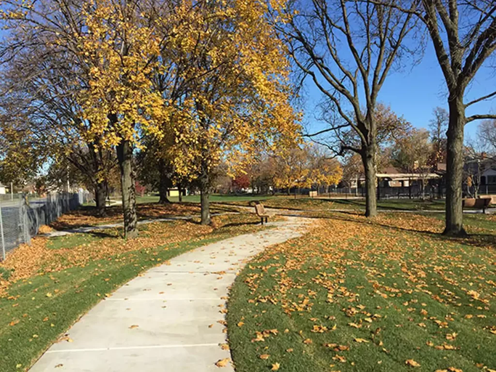 A paved walking path curves through a park in autumn, surrounded by trees with yellow leaves and fallen leaves on the grass. Benches are visible.