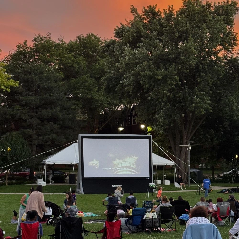 An outdoor movie screen is set up in a park at dusk, with an audience seated on the grass watching the film. A tent is