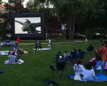 An outdoor movie screen shows a scene from a film. People sit on blankets and chairs on a grassy lawn watching the movie.