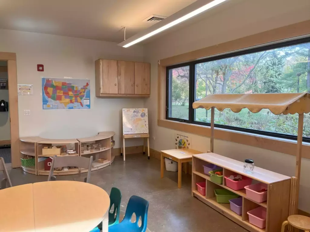 A bright, child-friendly classroom with a US map, an easel, a play market stall, and storage shelves filled with colorful bins.