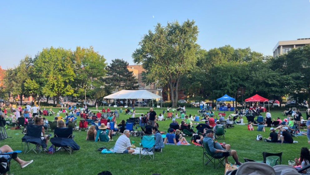 A large crowd sits on a grassy lawn watching a performance under a clear sky, with tents and trees in the background.