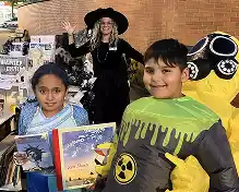 Two children in costumes, one holding books and the other in a hazmat suit, with a woman in a witch costume in the background at an event.