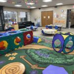 A colorful indoor playground area with a large modular climber, a pop-up tent, tables, chairs, and a nature-themed carpet.