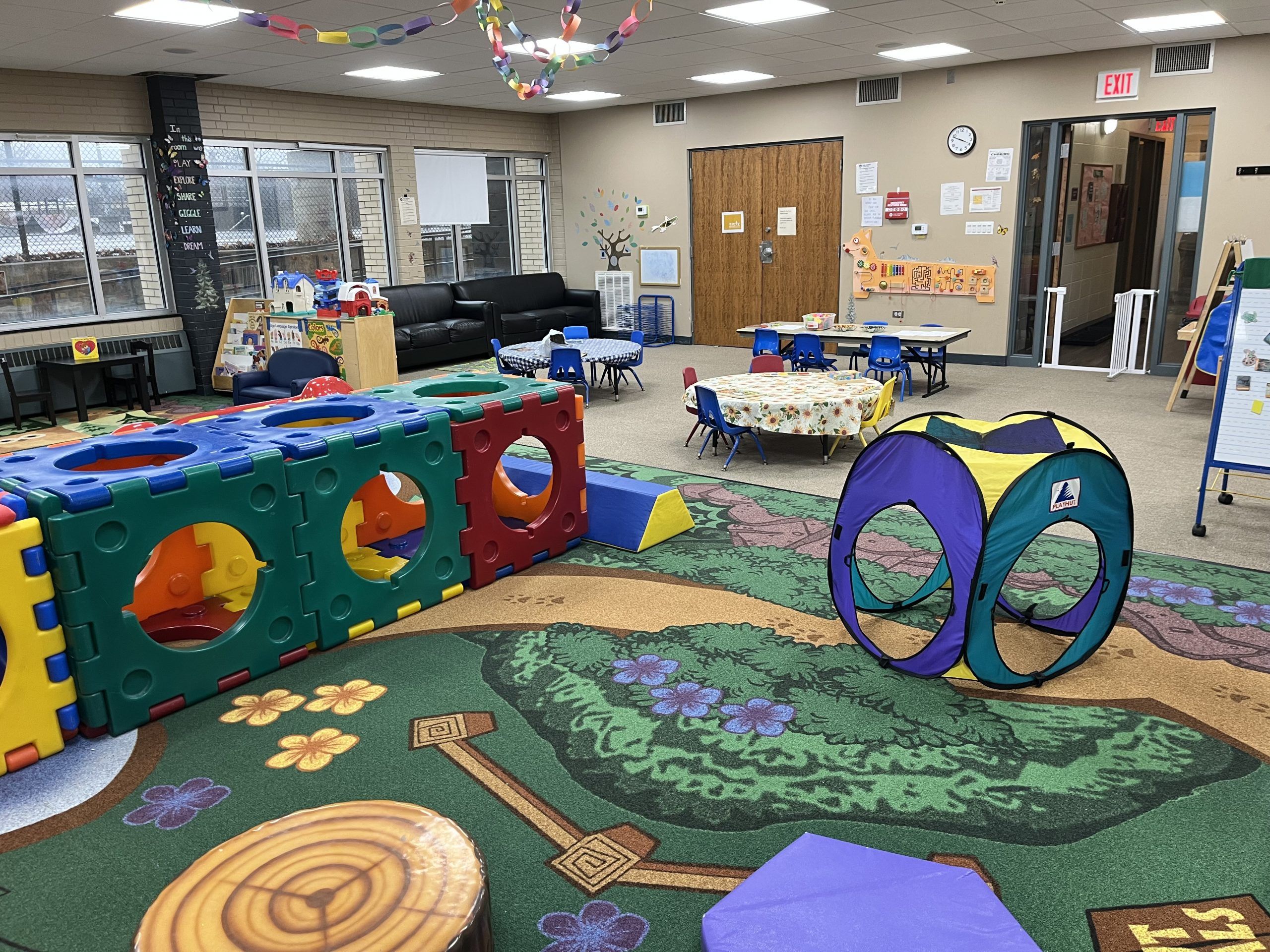 A colorful indoor playground area with a large modular climber, a pop-up tent, tables, chairs, and a nature-themed carpet.