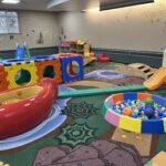 A colorful indoor playground featuring a large modular climbing structure, a red canoe-shaped seat, a ball pit, and slides, set on a nature-themed