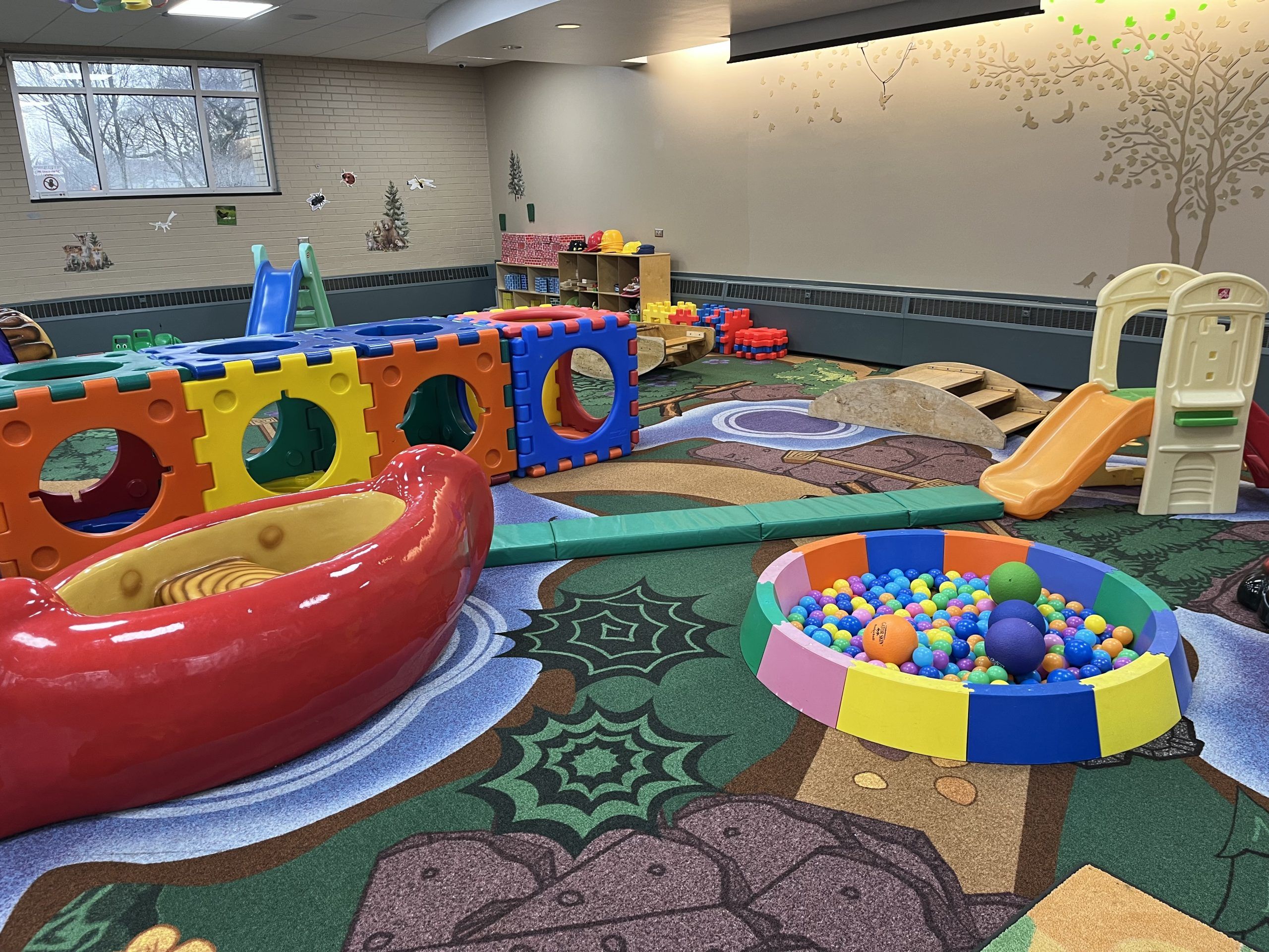 A colorful indoor playground featuring a large modular climbing structure, a red canoe-shaped seat, a ball pit, and slides, set on a nature-themed
