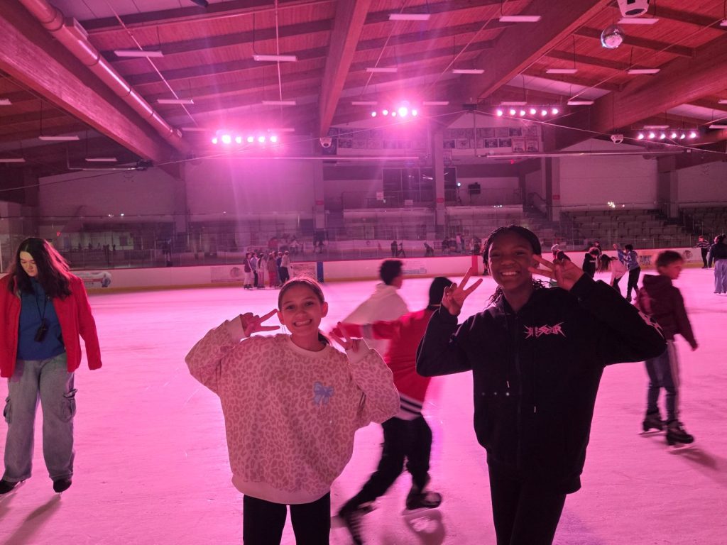 two skaters hold up peace signs for the camera with the rink lit in pink lights behind them