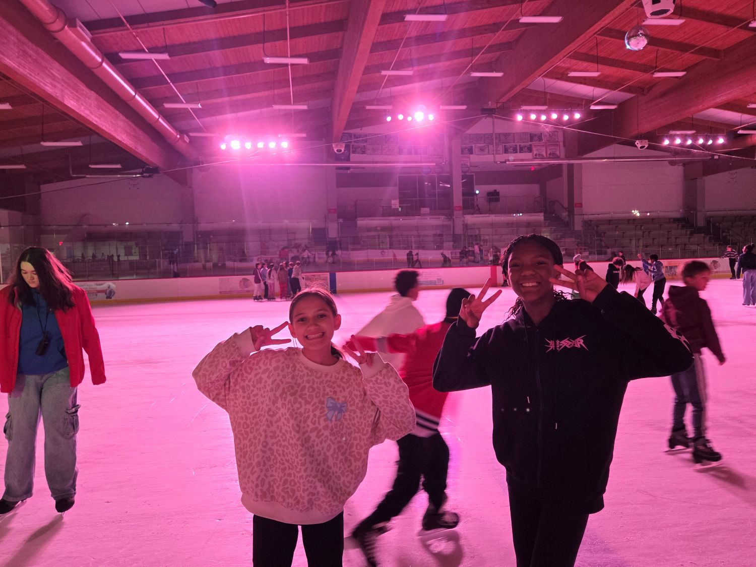 two skaters hold up peace signs for the camera with the rink lit in pink lights behind them