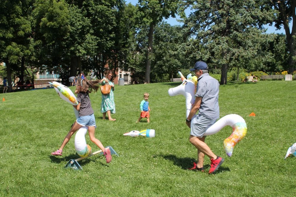 A girl and a man run with inflatable unicorns during an outdoor event. A young boy watches.